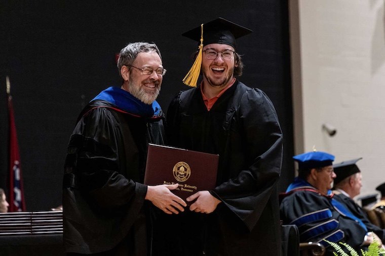 A graduate in a cap and gown smiles while holding a diploma, standing beside a faculty member in academic regalia at a graduation ceremony. Other faculty are seated in the background.