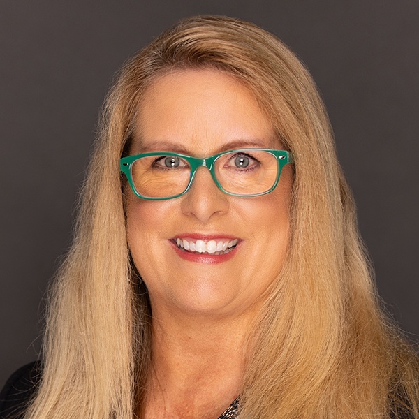 Smiling woman with long blonde hair wearing teal glasses and a black top, posing in front of a plain dark background.