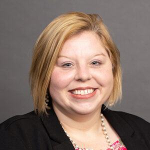 A woman with short blonde hair smiles at the camera. She is wearing a black blazer, a pink floral top, a pearl necklace, and pearl earrings. The background is plain and gray.