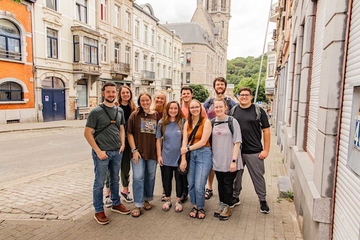 A group of ten young adults stands closely together, smiling for a photo on a cobblestone street lined with old buildings. A church with a tall tower is visible in the background.