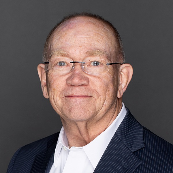 An older man with glasses and a receding hairline, wearing a dark pinstripe suit and white shirt, smiles slightly in front of a plain dark gray background.