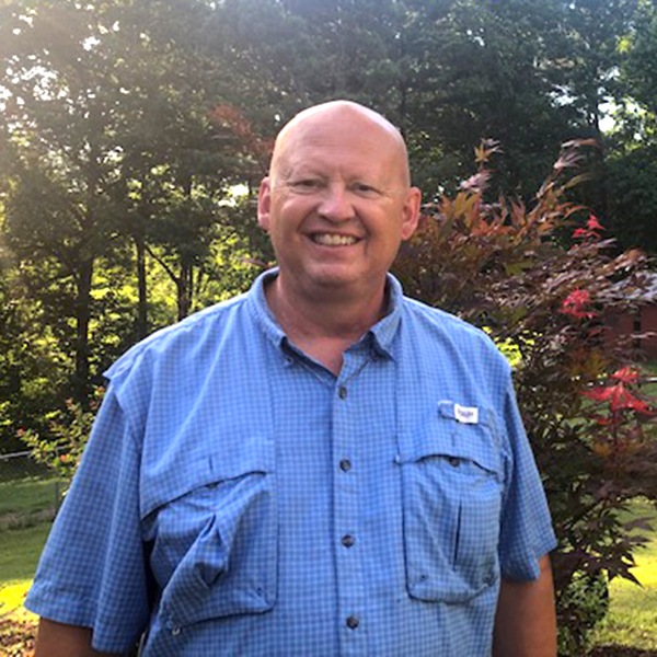 A smiling bald man wearing a blue short-sleeve button-up shirt stands outdoors in front of colorful foliage and trees, with sunlight filtering through the background.
