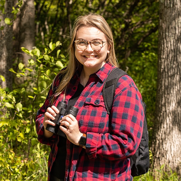 A smiling person with glasses wearing a red plaid shirt and backpack stands outdoors in a sunlit forest, holding binoculars.