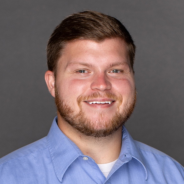 A man with short brown hair and a beard, wearing a light blue collared shirt, smiles in front of a plain dark gray background.