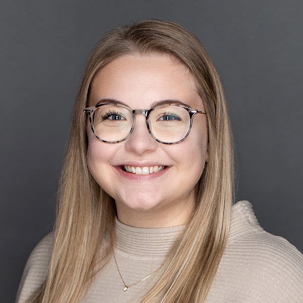 A young woman with long blonde hair, wearing round glasses and a beige top, smiles in front of a plain gray background.