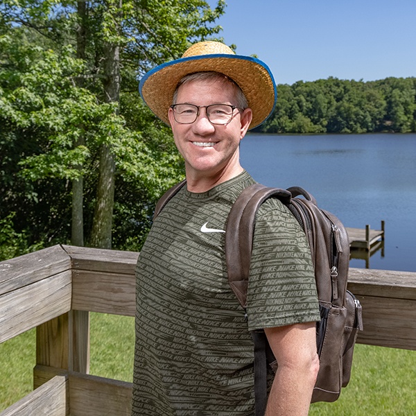 A smiling man wearing glasses, a straw hat, and a backpack stands on a wooden deck by a lake, surrounded by trees and greenery under a clear blue sky.
