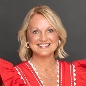 A smiling woman with short blonde hair wearing a red top with white trim, silver earrings, and layered necklaces, posing against a dark gray background.