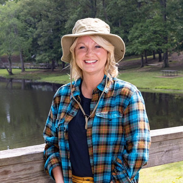 A smiling woman in a tan sun hat and blue plaid shirt stands by a wooden fence in front of a pond, with green trees and grass in the background on a sunny day.