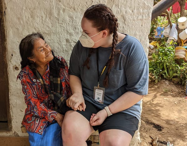 A young woman wearing a mask and name badge holds hands and smiles warmly with an elderly woman in colorful clothing, sitting together outside in a rural setting.