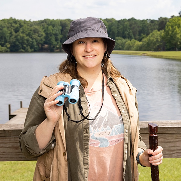 A woman in outdoor clothing and a bucket hat stands by a lake, holding blue binoculars and a walking stick, with trees and water in the background.