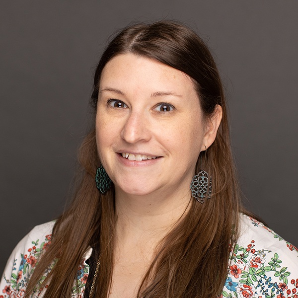 A woman with long brown hair smiles at the camera. She is wearing dark, floral-shaped earrings and a white top with colorful floral embroidery, set against a plain gray background.