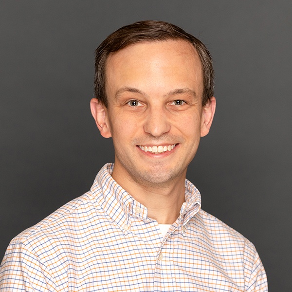 A man with short brown hair smiles at the camera, wearing a light-colored checkered shirt. The background is a plain, dark gray.