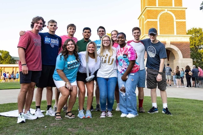 A group of twelve smiling young adults pose together outdoors on grass near a brick clock tower, wearing casual summer clothes and college T-shirts. Other people are visible in the background.