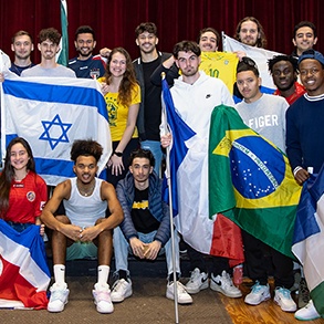 A diverse group of young adults poses indoors, smiling and holding flags from various countries, including Israel, Brazil, and France, against a red curtain background.