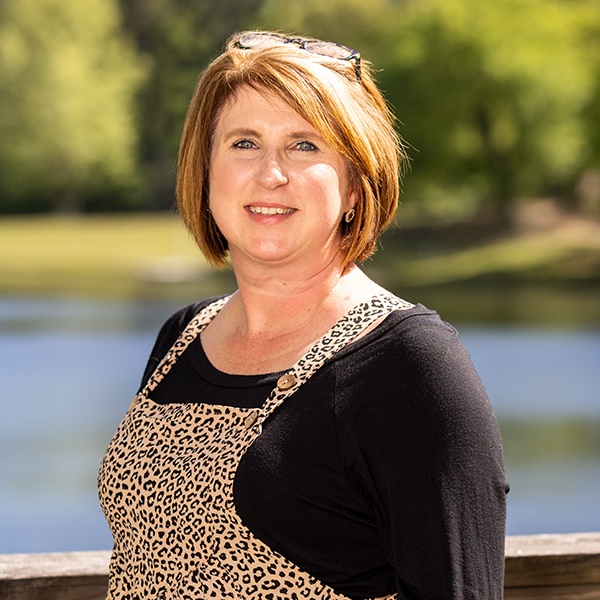 A woman with short, light brown hair stands outdoors in front of a lake and green trees, wearing a black shirt and leopard-print overalls, smiling at the camera. Sunglasses rest on her head.
