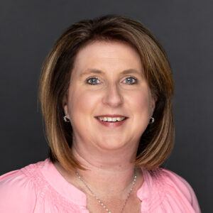 A woman with shoulder-length brown hair smiles at the camera. She is wearing a light pink top and small hoop earrings, with a dark gray background behind her.