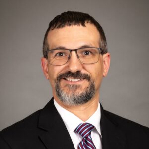 A man with short brown hair, glasses, and a beard is smiling at the camera. He is wearing a dark suit, a white shirt, and a striped tie. The background is plain gray.