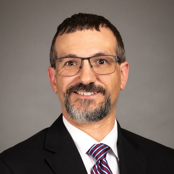 A man with short brown hair, glasses, and a beard is smiling at the camera. He is wearing a dark suit, a white shirt, and a striped tie. The background is plain gray.