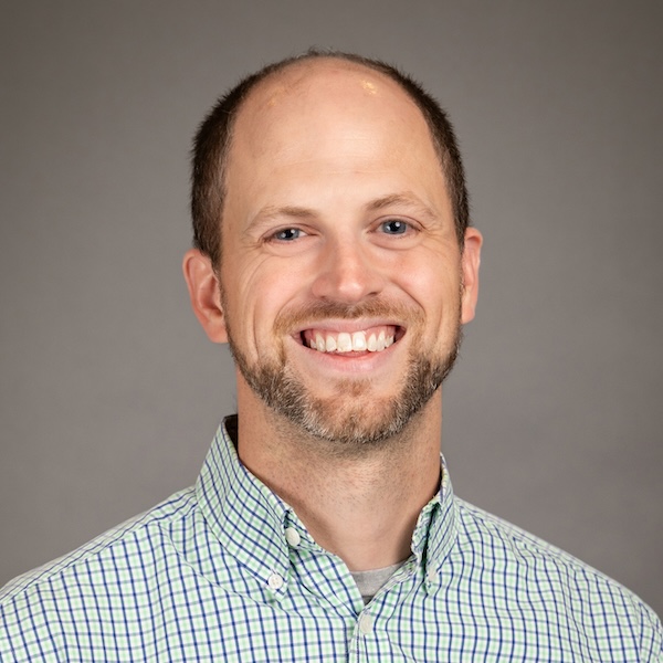 A smiling man with a short beard and receding hairline, wearing a light blue checkered shirt, posing in front of a plain dark gray background.