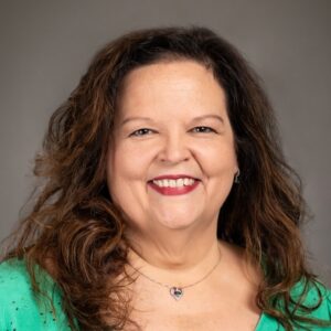 A woman with long brown hair smiles at the camera. She is wearing a colorful, patterned top and a necklace, with a neutral gray background behind her.