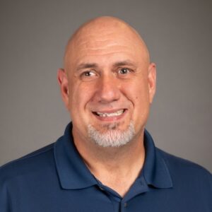 A middle-aged man with a bald head, trimmed goatee, and mustache, wearing a gray collared shirt, smiles slightly in front of a plain dark background.