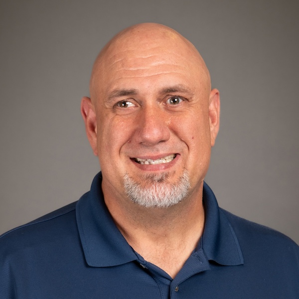 A middle-aged man with a bald head, trimmed goatee, and mustache, wearing a gray collared shirt, smiles slightly in front of a plain dark background.