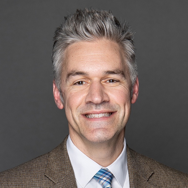 A smiling man with short gray hair wearing a brown suit jacket, white dress shirt, and a blue plaid tie, posed against a plain gray background.