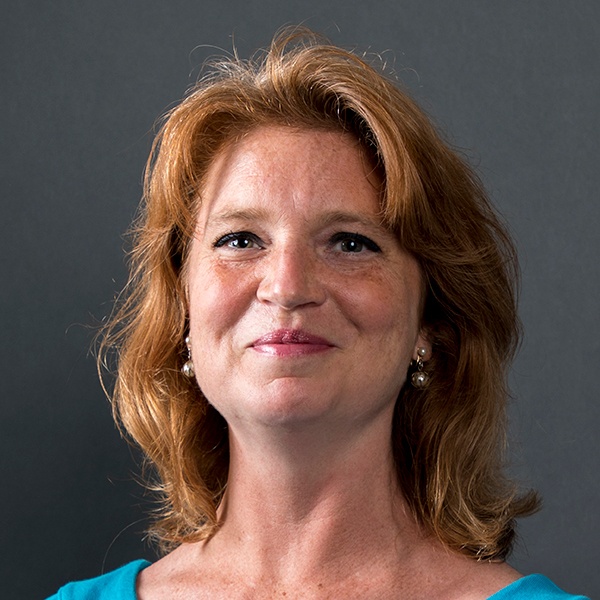 A woman with shoulder-length reddish-brown hair and light skin, wearing pearl earrings and a turquoise top, smiles slightly in front of a plain dark gray background.