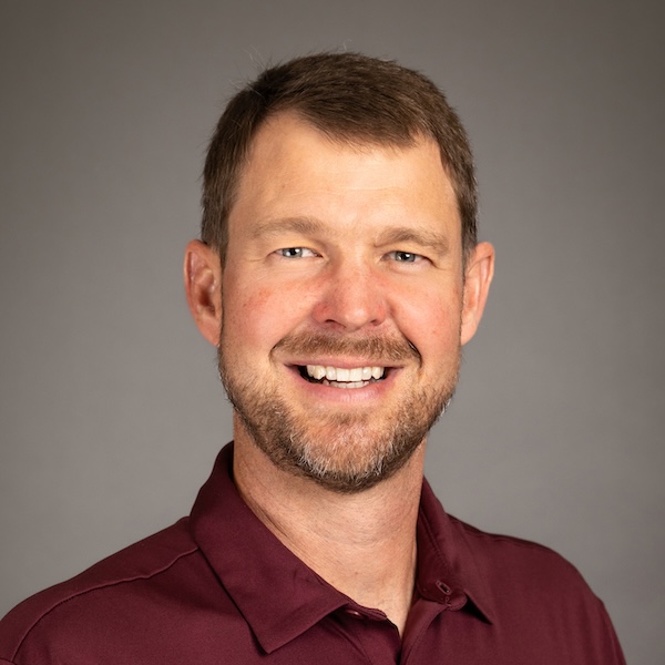 A man in a navy suit, light blue shirt, and red tie is smiling in front of a plain gray background. He has short brown hair and a trimmed beard.
