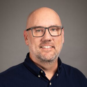 A middle-aged man with glasses, a light beard, and short hair, wearing a navy blazer and a light blue checkered shirt, smiling against a plain gray background.