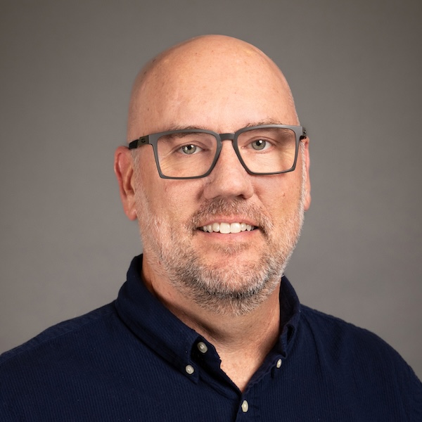 A middle-aged man with glasses, a light beard, and short hair, wearing a navy blazer and a light blue checkered shirt, smiling against a plain gray background.