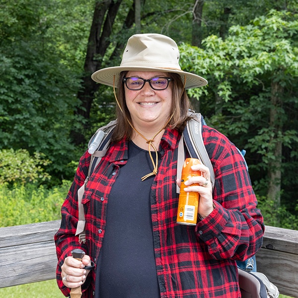 A smiling woman wearing glasses, a wide-brimmed hat, and a red plaid shirt holds a spray bottle and a hiking stick while standing outdoors in a wooded area.