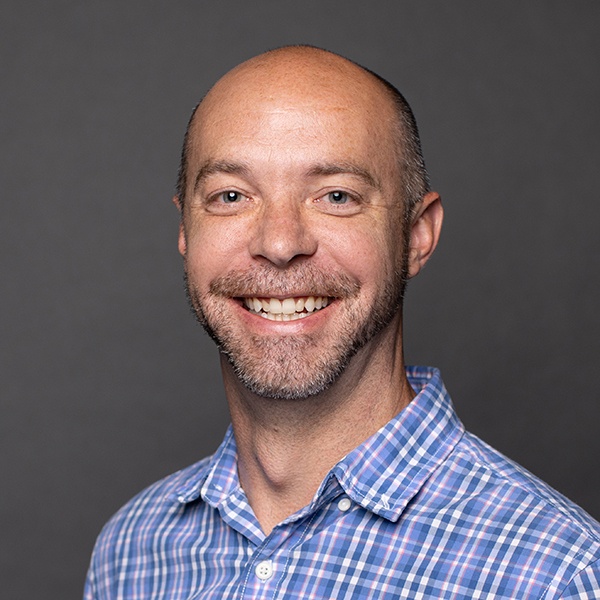 Smiling man with a bald head and short beard wearing a blue and pink plaid shirt, standing against a plain gray background.