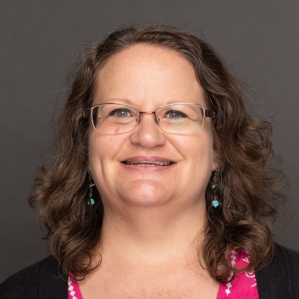 A woman with wavy brown hair and glasses smiles at the camera. She is wearing a pink top with a patterned neckline, turquoise earrings, and is set against a plain dark background.