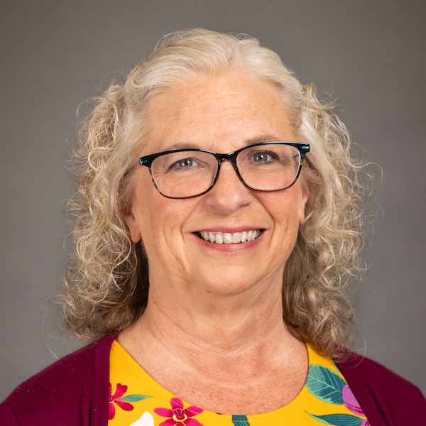 Smiling older woman with curly gray hair, wearing glasses, a black top, a light gray cardigan, and a round pendant necklace, posing in front of a plain gray background.