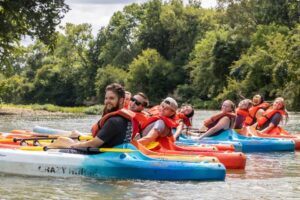 A group of eight people wearing life jackets smile and pose for a photo while sitting in colorful kayaks on a river, surrounded by trees and greenery on a sunny day.