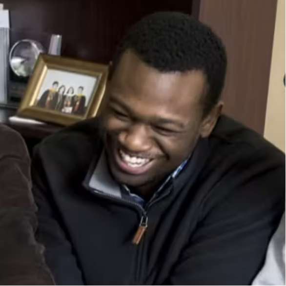 A man with short hair and a black jacket is smiling and looking down. In the background, there is a wooden shelf with a framed group photo and other decorative items.