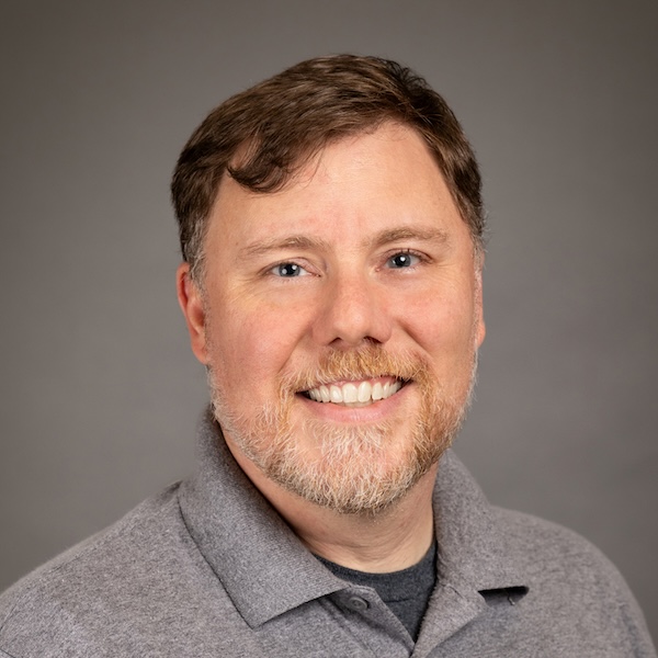 A smiling man with short light brown hair and a beard, wearing a light green polo shirt, posing in front of a plain gray background.