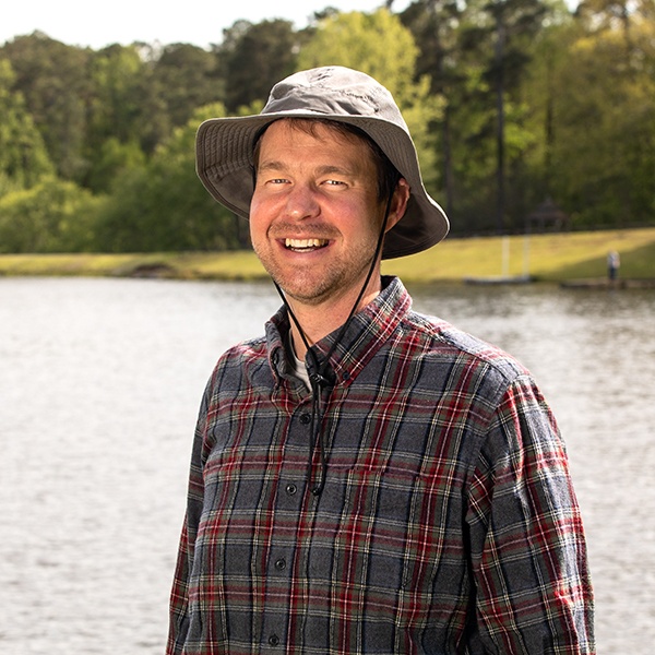 A smiling man in a plaid shirt and wide-brimmed hat stands by a lake with trees and greenery in the background, enjoying a sunny day outdoors.