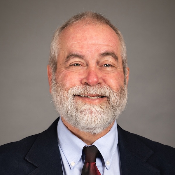 A middle-aged man with a gray beard and short hair smiles, wearing a dark suit, patterned tie, and checkered shirt, posing in front of a plain gray background.