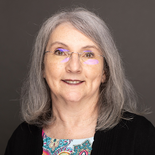 Smiling older woman with long gray hair and glasses, wearing a black sweater over a colorful patterned top, in front of a plain dark background.