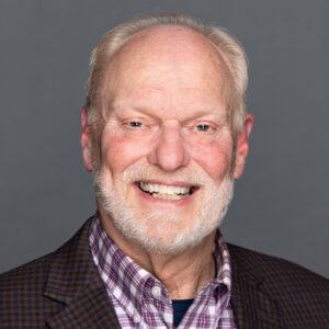 Smiling older man with white hair and beard wearing a plaid shirt and checked blazer, posed in front of a plain gray background.