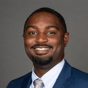 A man in a blue suit and patterned tie smiles at the camera against a plain gray background. He has short hair, a trimmed beard, and is wearing a light blue shirt.