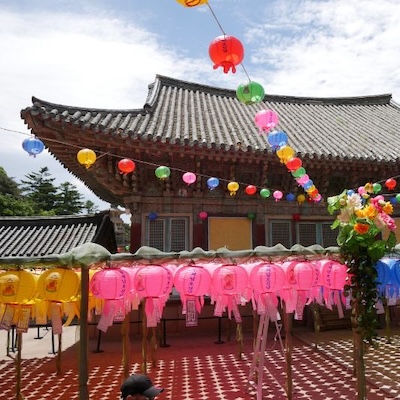 Colorful paper lanterns hanging across the courtyard of a traditional Korean temple building under a blue sky.