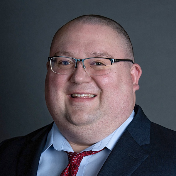 A smiling person with glasses, wearing a suit, light blue shirt, and red patterned tie, stands in front of a plain dark background.