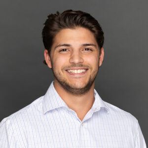 A young man with short brown hair and a beard smiles at the camera. He is wearing a white, collared, checkered shirt and is posed in front of a plain gray background.