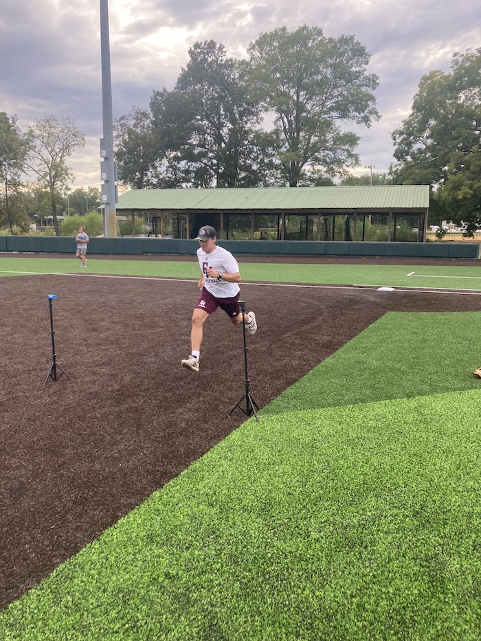 A person in athletic gear runs on a baseball field, passing between two timing sensors. Trees, a fence, and a covered seating area are visible in the background under a cloudy sky.