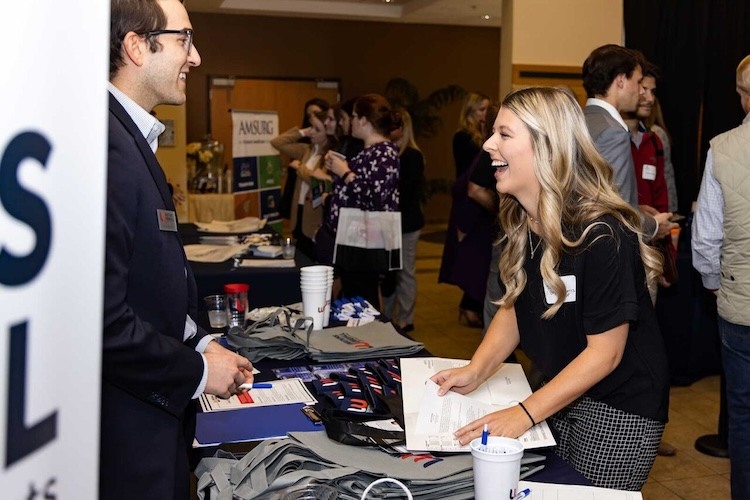 A woman smiles and leans over a table while talking to a man at a busy indoor event. Both are wearing name tags, and promotional materials and tote bags are spread out on the table between them.