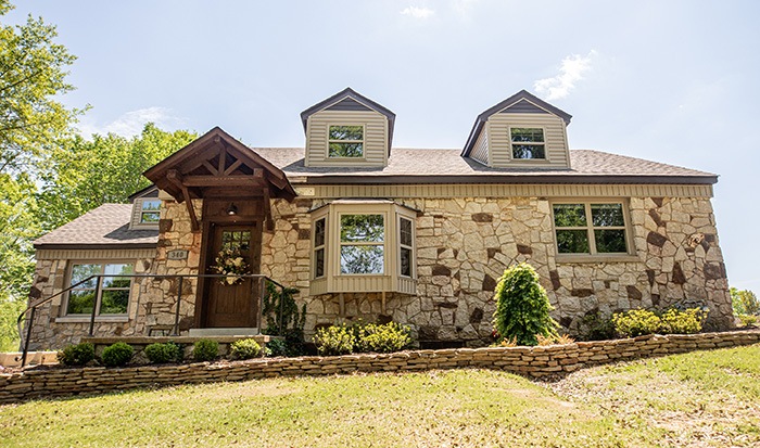 A stone house with two dormer windows, a wooden front door with a peaked porch, and landscaping featuring bushes and a small tree in front, set on a slightly sloped lawn under a sunny sky.