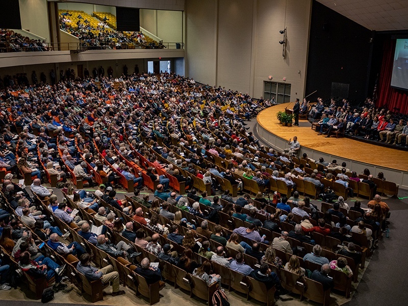 A large audience fills an auditorium with rows of seats, watching a presentation on stage where several people are seated and one person is speaking at a podium. The upper balcony is also crowded.